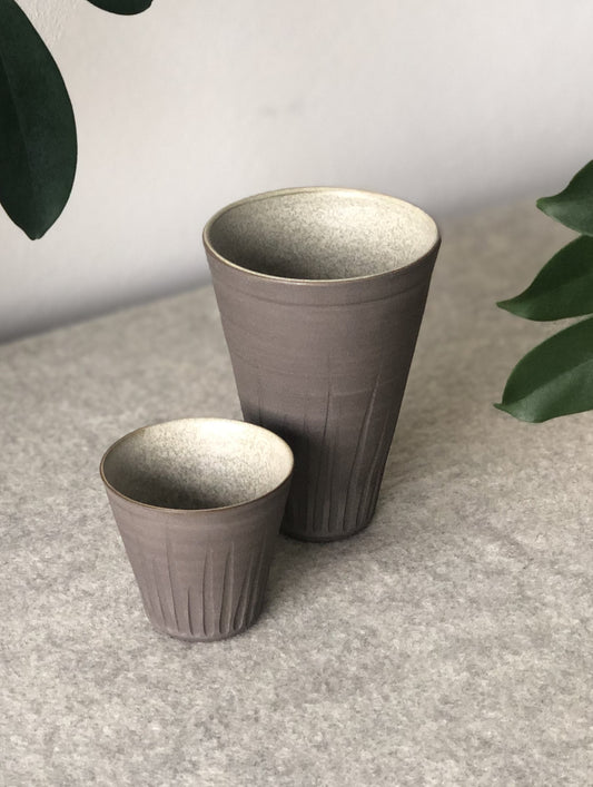 Photo of a grey coloured clay beaker next to a grey coloured espresso pot on a light grey fabric material..