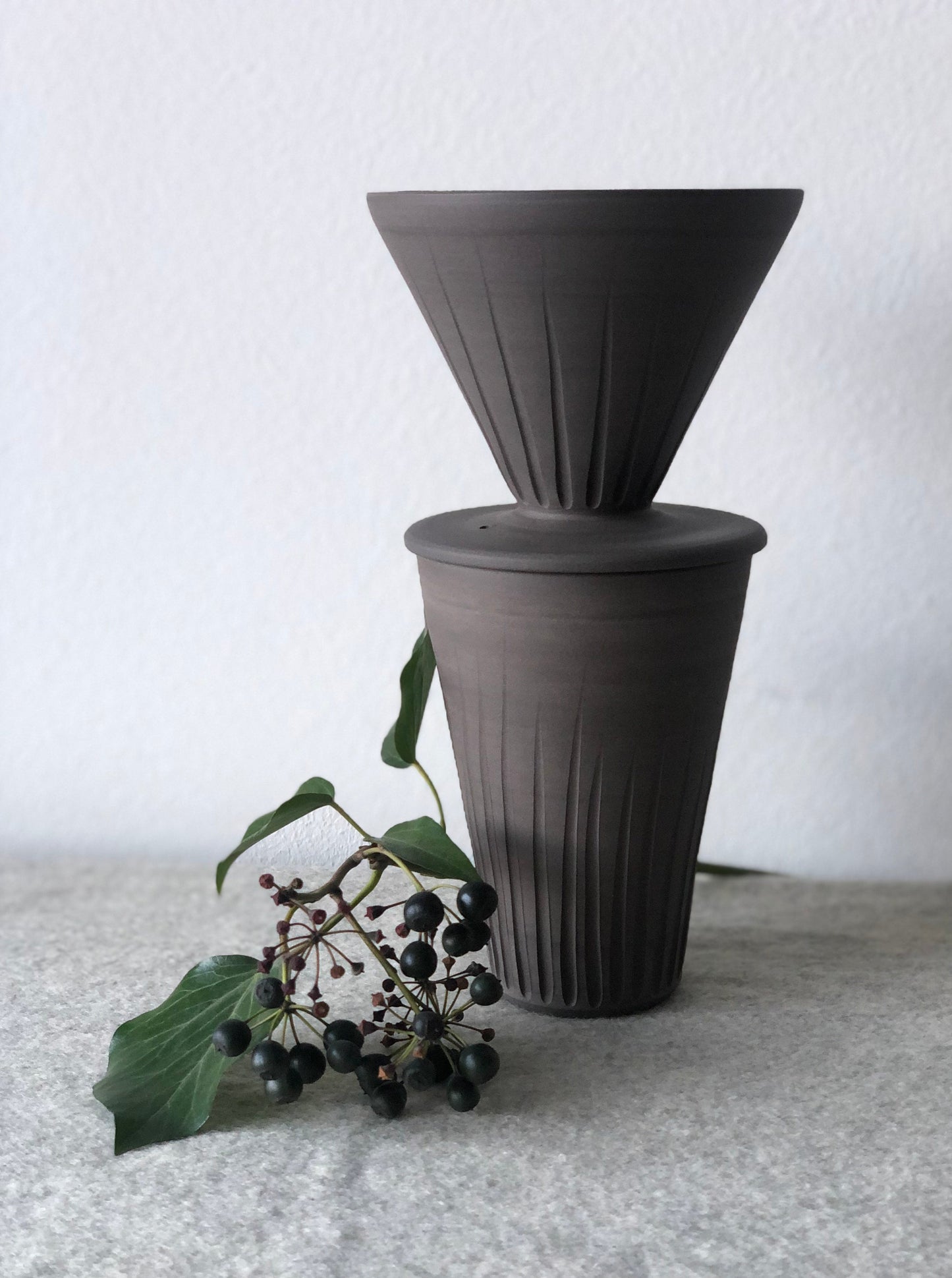 Charcoal coloured pottery coffee ourover stancked onto of a straight sided tall beaker. Small berry branch decorating the base of the pots.